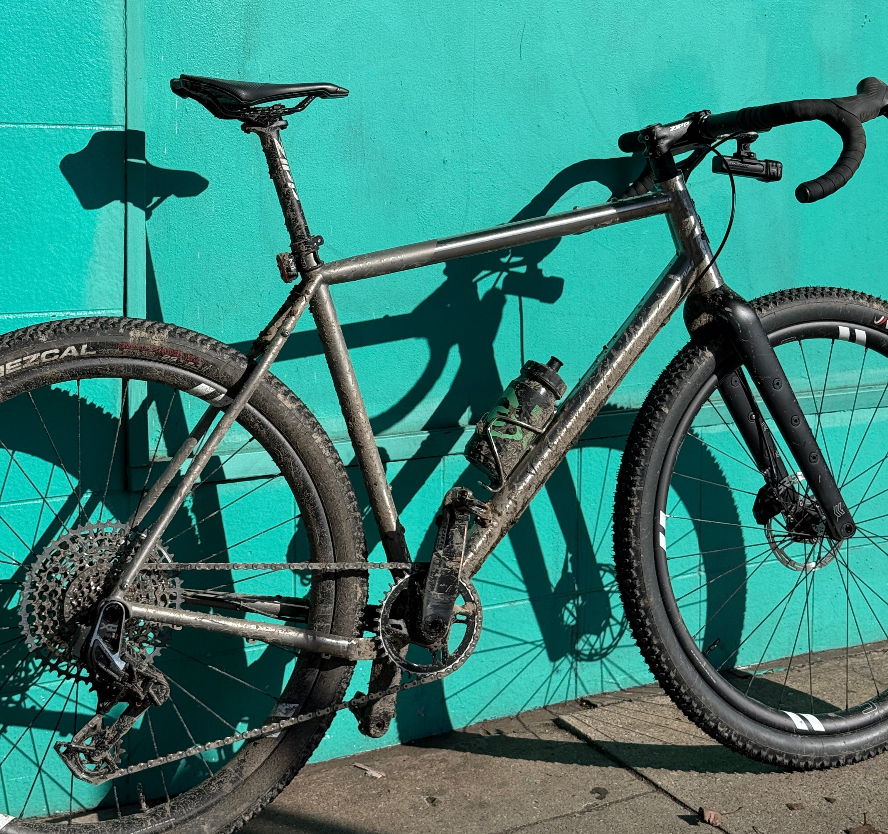 A used mountain bike with a dark gray frame and black components, lying on a bright turquoise tiled floor next to a concrete wall.